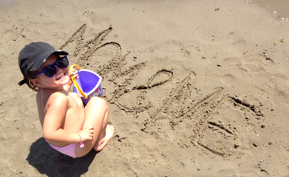bambina mom&me momeme scritta sulla sabbia bagnata al mare con cappello e secchiello